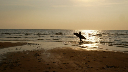 冲浪者在热带海滩日落时带着长的冲浪板踏入海中视频