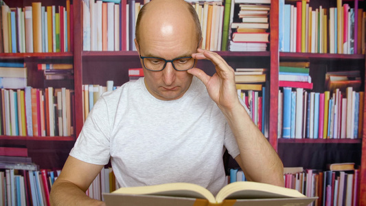 在家庭办公室桌边看书的男用眼镜的人以书架背景视频