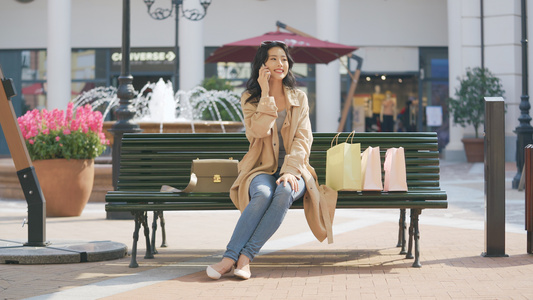 4k女性购物休息坐在长椅上使用手机打电话视频