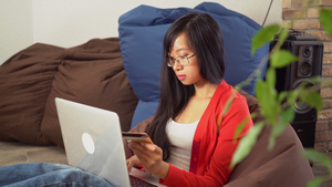 年轻的亚洲女人在坐在豆袋椅上的电脑上在线购买15秒视频