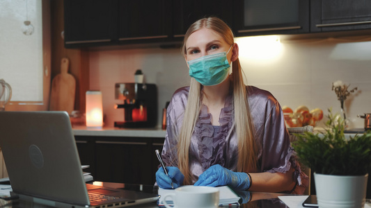 身戴医疗面具和手套的金发妇女在家工作对监视器微笑视频