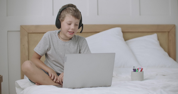 小男孩坐在家里的床上用笔记本在网上聊天用耳机麦克风视频