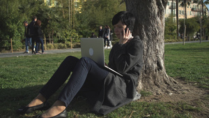女人在笔记本电脑上打字在公园的树下用智能手机说话13秒视频