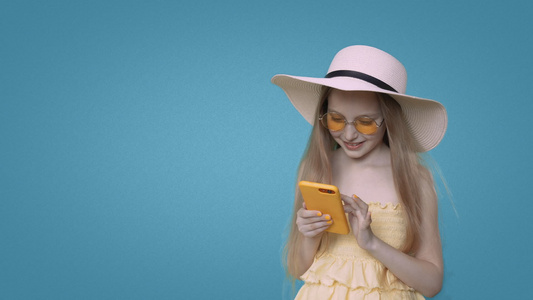穿着黄色衣服的笑着女孩在蓝背景上浏览智能手机视频
