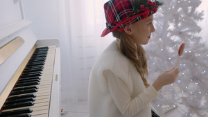 戴红帽的小女孩在白钢琴和圣诞树旁舔棒棒糖15秒视频