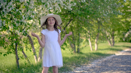 快乐的女孩戴着白帽子穿着春天花园里开花的树木背景在视频