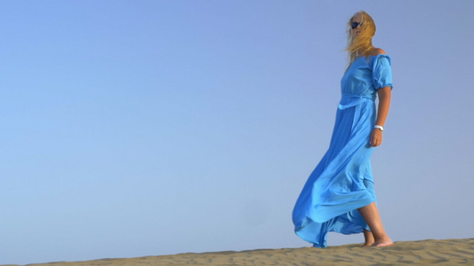 年轻金发女子身着蓝色长裙站在蓝天的慢动作视图它在风中视频