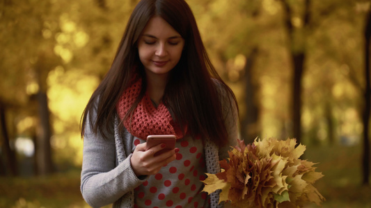 秋天女孩在公园散步在她的智能手机上使用应用程序录像片视频