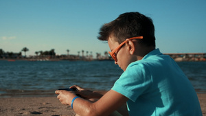 穿着蓝色T恤的小男孩坐在海湾拿手机打游戏20秒视频