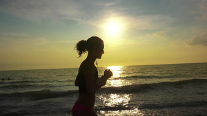 穿着马尾和耳机的跑漂亮女孩在夏季海滩日落时放松慢跑16秒视频