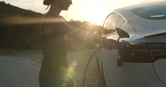 女孩在日落时用电动汽车插上充电电缆手持录像视频