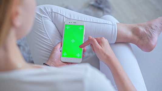 穿着白色牛仔裤坐在沙发上的年轻妇女使用智能手机加绿屏视频