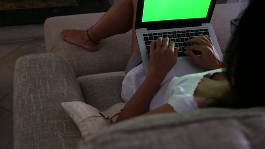 一名年轻妇女在她的笔记本电脑上工作,在家用绿色屏幕做画,坐在她客厅的沙发上视频