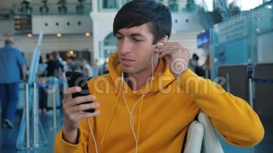 坐在机场休息室的长椅上，男人一边看着智能手机屏幕，一边通过耳机听音乐视频