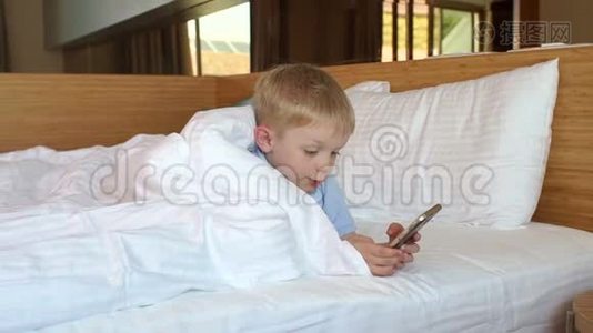 一个在家打电话的小男孩躺在毯子下面的床上。视频