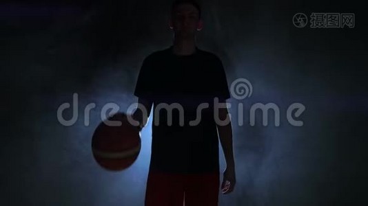 穿着红色短裤和蓝色t恤的篮球运动员在一个黑暗的篮球场，背光即将到来视频