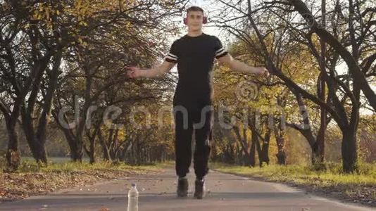 日落时分，一位戴着耳机和黑色运动服的高加索青年运动员在秋天公园跳绳的肖像视频