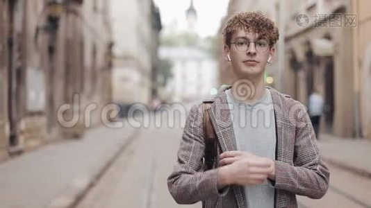 戴眼镜和戴耳机的年轻微笑的嬉皮士男子站在旧城街道背景下的画像视频