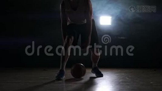 一个年轻的成年人，篮球运动员运球，黑暗的室内篮球场。 慢烟视频
