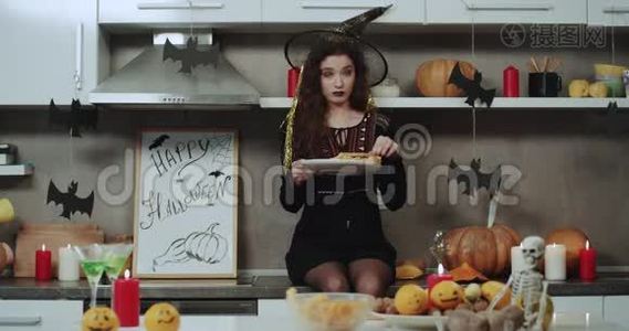 一个年轻的蒙面女巫坐在厨房的桌子上，拿着一个小吃盘在镜头前玩。视频