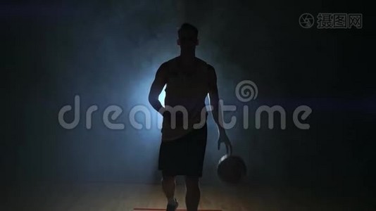 篮球运动员在一个黑暗的房间里径直走向摄像机，烟雾中有一个背光的背部看着摄像机。视频
