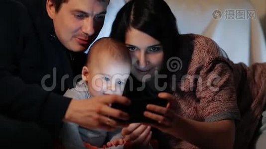 年轻快乐的父母和他们的小儿子看着智能手机屏幕微笑。 慢镜头。视频
