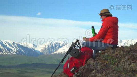 旅行者坐在山顶的女人，从热水瓶里喝热茶。 红色背包，橙色夹克。 草帽。 筷子视频