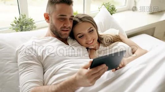年轻可爱的一对情侣，早上在卧室里躺在床上用平板电脑聊天视频