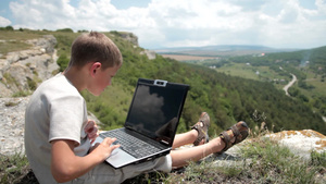 男孩在山上悬崖边缘的笔记本电脑上工作21秒视频