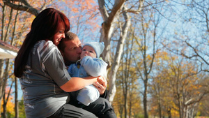 秋天一家三口坐在公园的椅子上父亲在亲吻妈妈怀里抱着的婴儿10秒视频