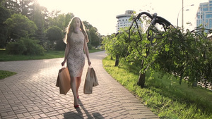 夕阳下时尚的金发女人穿着红色高跟鞋和连衣裙提着购物袋走在城市街道上17秒视频