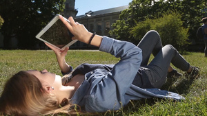 漂亮的金发女孩躺在绿色的草地上用触控板在校园里学习特写镜头14秒视频