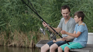 快乐的父亲和他的十几岁的在夏天坐在木墩上拿着鱼竿在湖边钓鱼和聊天30秒视频