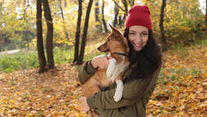 快乐积极的年轻女人在帕卡和红色针织帽抱着她可爱的狗微笑着在金色的秋天景观背景13秒视频