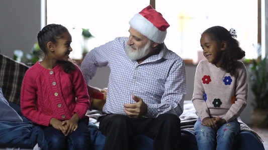 快乐的祖父戴着圣诞帽扮演圣诞老人给两个混血孙女送圣诞礼物视频