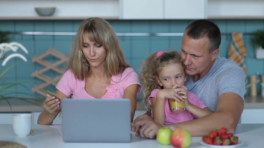 美丽的父母和他们的小女儿早上一起坐在厨房里用笔记本电脑和信用卡购物视频