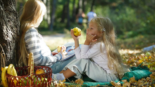 两个金发小女孩坐在大树下享受阳光明媚的秋天在看书和吃苹果视频