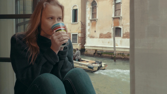 年轻女子喝着热咖啡坐在威尼斯运河旁的窗台上视频