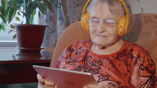 老年妇女在家里坐在扶手椅上用平板电脑和无线耳机听音乐视频