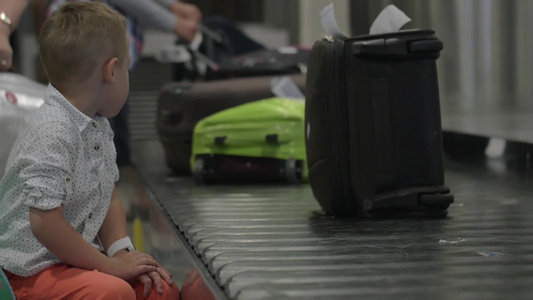 一个小男孩坐在机场的行李传送带旁看着人们拿行李的慢动作视频