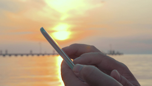 在美丽日落的湖边一位男子手里拿着智能手机的特写镜头视频