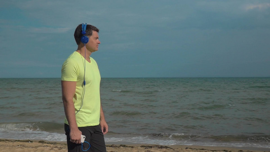 一个年轻人站在海滩上手里拿着智能手机戴着耳机听着音乐看望远方的人视频