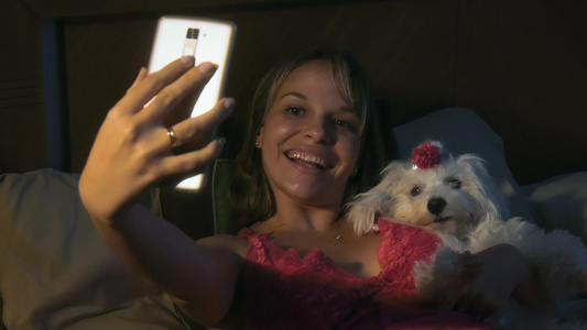 年轻的女人晚上拥抱她的小狗用手机拍自拍视频