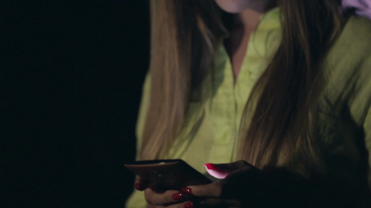 在户外年轻漂亮女人在夜间模糊的灯光背景下使用智能手机上发短信视频