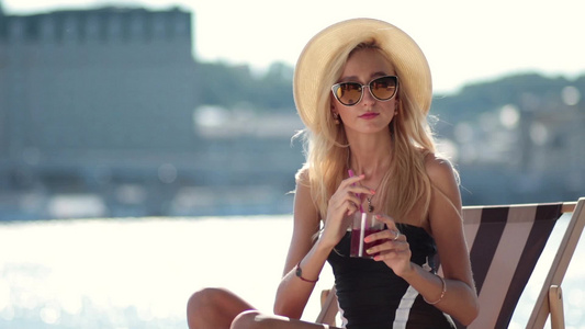 戴着帽子和太阳镜的金色长发美女坐在海滩上的太阳躺椅上喝鸡尾酒视频