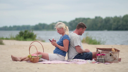 中年夫妇背靠背坐海滩上野餐玩手机视频
