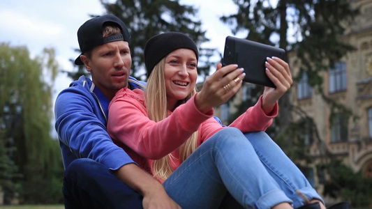 快乐的一对学生坐在大学外的公园的草地上与他们的朋友在线使用电子数字平板电脑进行视频交流视频