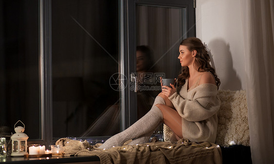 冬天,舒适人的年轻的女人穿着套衫,坐窗台上,家里喝咖啡或茶杯家里窗台上有咖啡或茶杯的女人图片