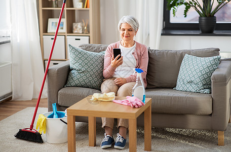 家庭技术快乐的高级妇女用智能手机后,清洁家庭老年妇女打扫家后用智能手机图片