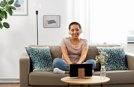 人,技术休闲快乐微笑的年轻非裔美国妇女戴着耳机,坐沙发上,家听音乐或平板电脑上看电影家里有耳机平板电脑的女人图片